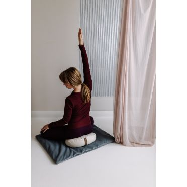 Kobieta w bordowym stroju siedzi z wyprostowaną ręką na jasnej poduszce fasolce umieszczonej na ciemnoszarej macie.